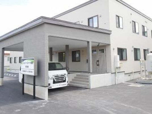 グレー系の色でまとめられた2階建ての建物の玄関には手すり付きのスロープと階段があり、車が止められている。