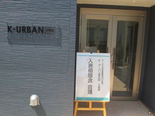 グレーの外壁には黒い文字で住宅名が書かれたプレートが貼られ、玄関の前には相談会と書かれた看板が立っている。