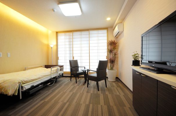 居室例。ベッドとイス、大きな液晶テレビが置いてある。間接照明や、大きな窓から光が入り、あかるい空間