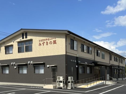 ベージュとダークグレーの２階建ての建物の外壁にはホーム名が書かれ、玄関先には手すり付きのスロープが付いている。