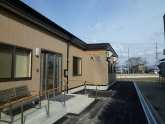 ブラウン系の建物は平屋建てで玄関には手すり付きのスロープがあり、ベンチも置かれている。
