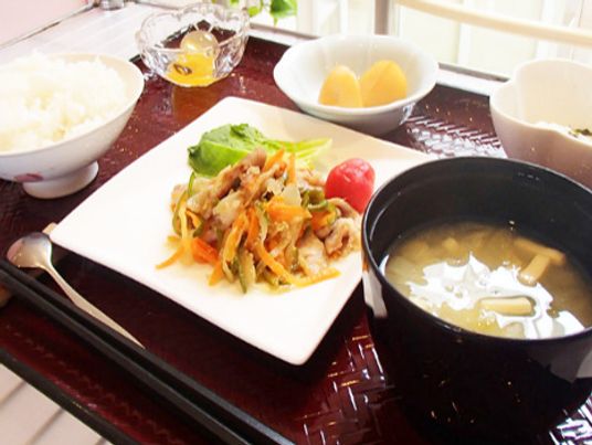 野菜炒めや白いご飯、みそ汁、野菜の小鉢にフルーツなどがそれぞれ皿に盛られている。