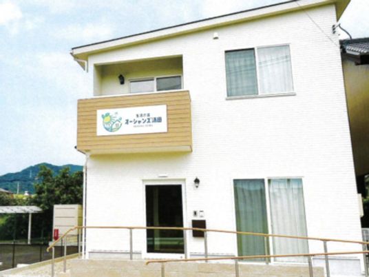 白い外壁の２階建ての建物は日当たりがよく、２階部分にベランダがあり、玄関には手すり付きのスロープがある。