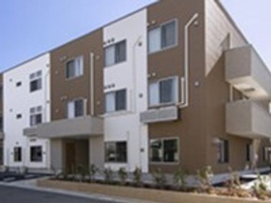 白とブラウンの3階建ての建物は角フロアにベランダが付いている。玄関には屋根があり、駐車スペースもある。