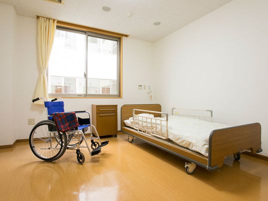 車いすや寝具付きのベッドが置かれた部屋にはカーテンが付いた大きな窓がある。