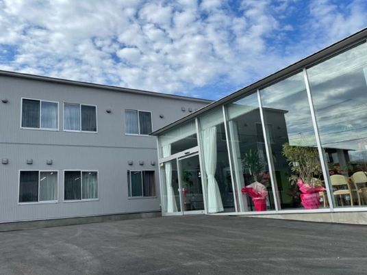 2階建ての白っぽい外壁の建物が左側にあり、右側には一面ガラス窓の平屋建ての建物があり、館内にテーブルセットや観葉植物がある。