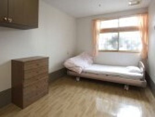 介護ベッドやタンスが置かれたフローリングの部屋には、カーテンが付いた大きな窓がある。