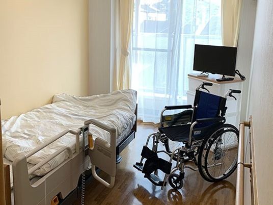 寝具付きのベッドが置かれ、車いすやテレビが置かれている部屋にはレースカーテンとカーテンが付いた大きな窓がある。