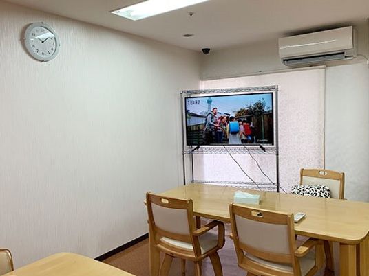 テレビが設置されている食堂には木製のダイニングテーブルが複数置かれ、時計やエアコンもある。