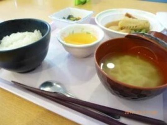 みそ汁に白いご飯、煮物、青菜の和え物などが並んでいるトレーには箸とスプーンがある。