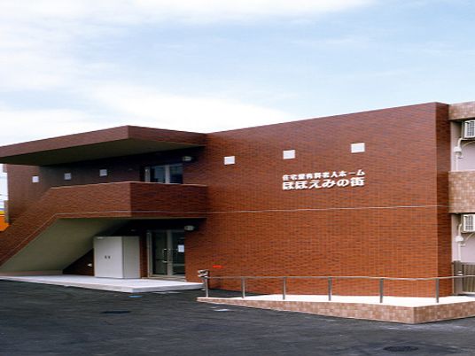 ブラウン系の2階建ての建物の外壁にはホーム名が書かれ、玄関先には手すり付きのスロープが付いている。