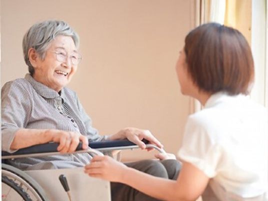 眼鏡をかけた高齢女性が車いすに座り、笑顔で白衣を着た女性スタッフと話している。