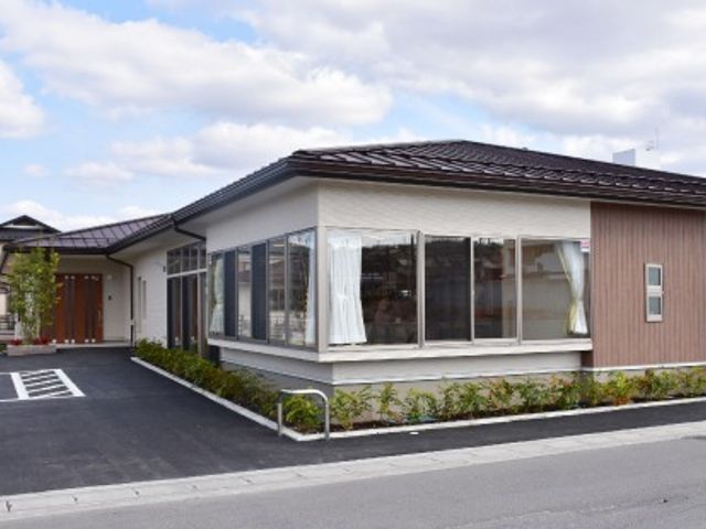 平屋建ての建物は落ち着いた色合い。駐車場が付き、建物の周りには木が植えられている。 ご利用される方々のニーズに多様な施設とサービスで応える「マインド」、スタッフの心が当グループの１番の魅力です。