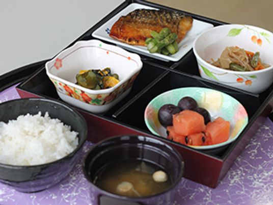 サバの照り焼きにフルーツ、野菜の小鉢が二品4月に区切られた弁当箱風の入れ物に入れられ、白いご飯と汁物が添えられている。