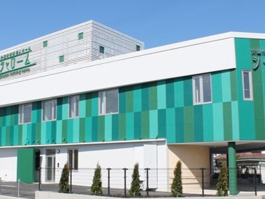 白とグリーンの建物は2階建てで日当たりのいい場所に建っていて、建物の周りには草木が植えられている。