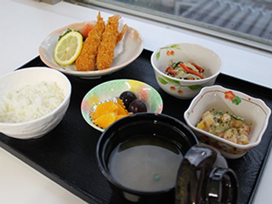 エビフライや野菜の和え物、煮物、白いご飯、みそ汁、フルーツが並んでいる。