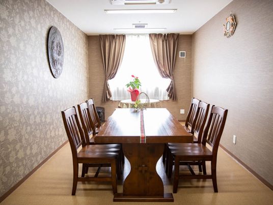 談話室に、木製の椅子が６脚とテーブルがある。窓際の丸テーブルの上には鉢に植えられた植物が飾られ、赤色の花が咲いている。