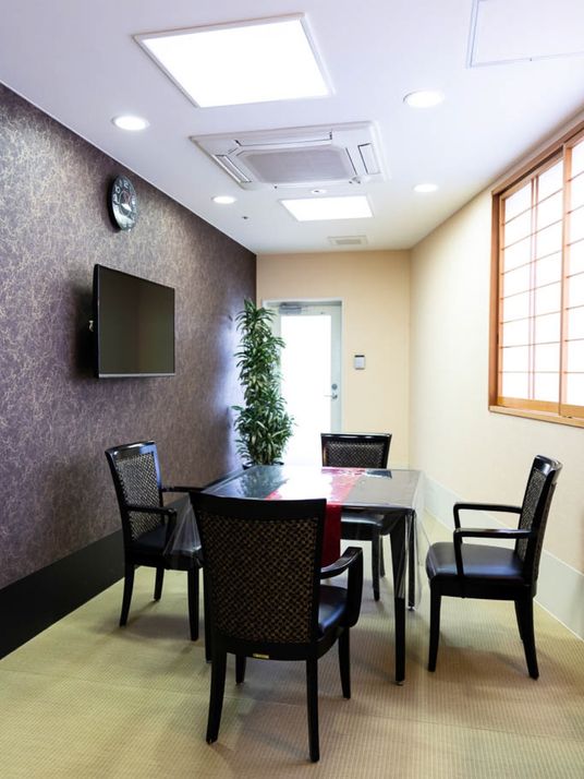 談話室には、黒色のテーブルと４脚の椅子が置かれている。壁掛けテレビが設置されている。背の高い観葉植物が置かれている。