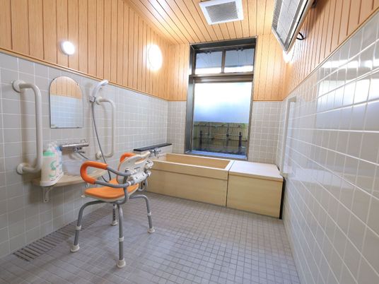 オレンジ色のシャワーチェアが備わっている。シャワーの両サイドには手すりがついている。シャンプーやリンスが用意されている。