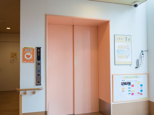 ピンクの扉と掲示物 エレベーター ゆとりある空間のエレベーター前です。AEDを含む安全設備も整い、閲覧しやすい掲示物があります。