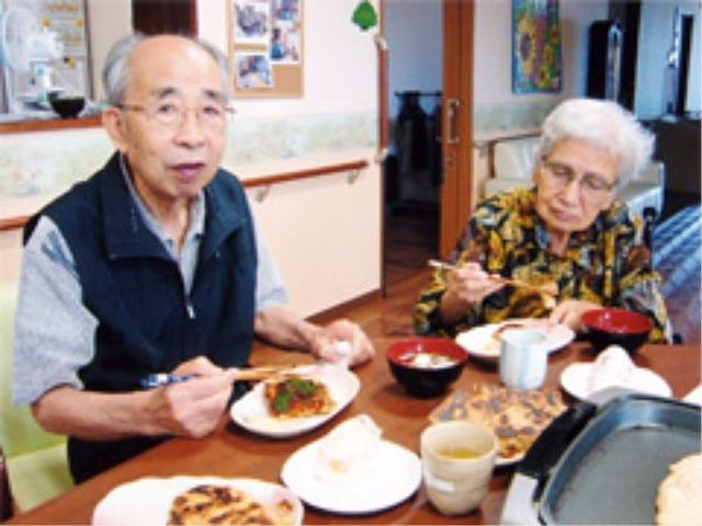 食事をとる高齢者ペア 食堂 落ち着いた雰囲気の中で、美味しそうな食事を楽しんでいる利用者様がおられます。ここの環境は、食事の時間も温かい交流の場となっています。