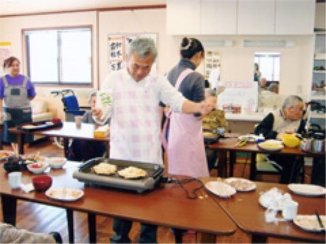 調理活動する人々の居室 レクリエーション 共同で調理を行なっている清潔感のあるダイニングルームです。スタッフと利用者が協力し合いながら、和気あいあいとした雰囲気の中で食事の支度をしています。