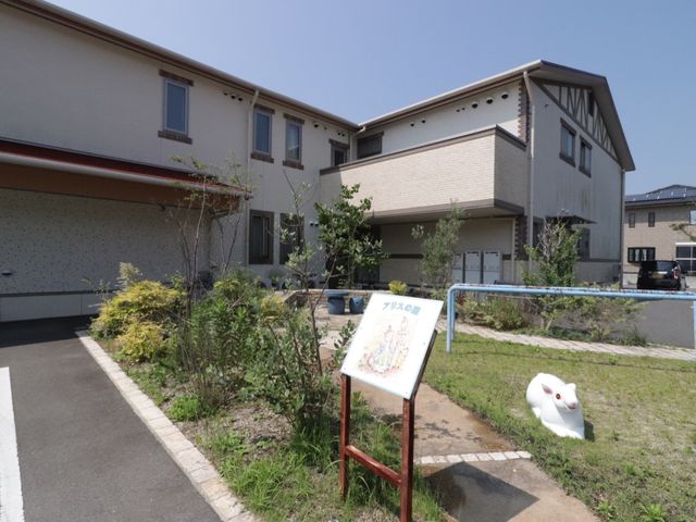明るい日差しの庭と建物 施設表示板 庭には植物が生い茂り、柔らかな光を浴びる建物が明るく迎えます。快適な環境でのお仕事をご検討ください。