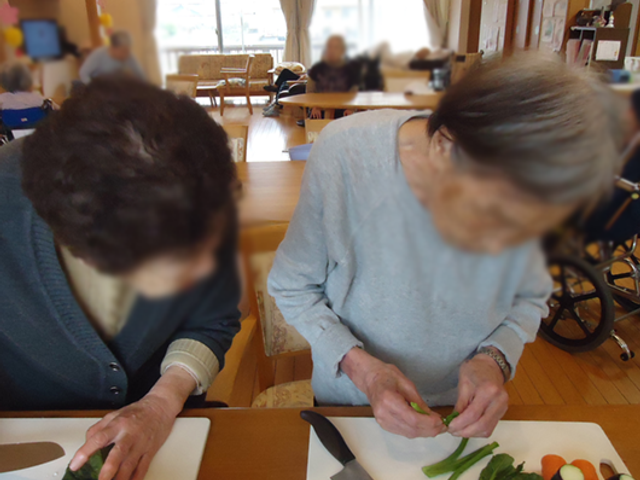 調理風景の共有スペース リビング 広々とした明るい空間で、入居者同士の交流や活動が行われています。料理を楽しむ姿も見られます。