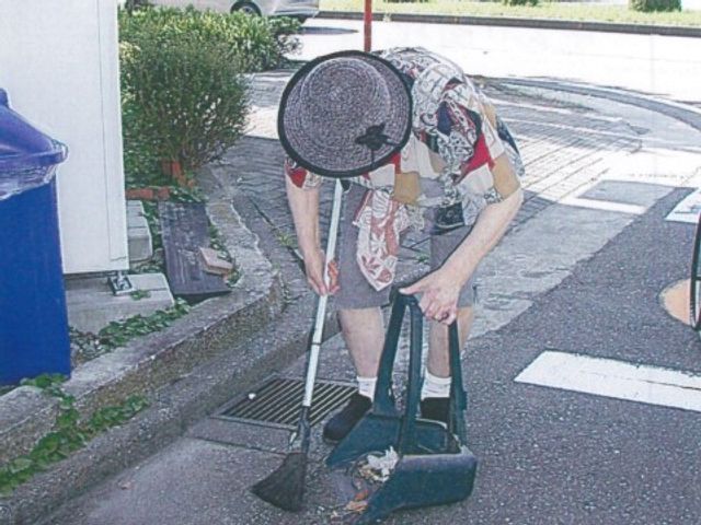 清掃を行う利用者様外観 入居者様 利用者様が杖をつきながら外で清掃活動をされており、自立した日常生活をサポートする環境が整っています。