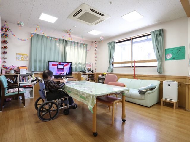居室の明るい一角 談話室 家庭的な雰囲気の居室で、テーブルを囲んだ共同生活が送れます。ソファやテレビがあり、リラックスできるスペースが備わっています。