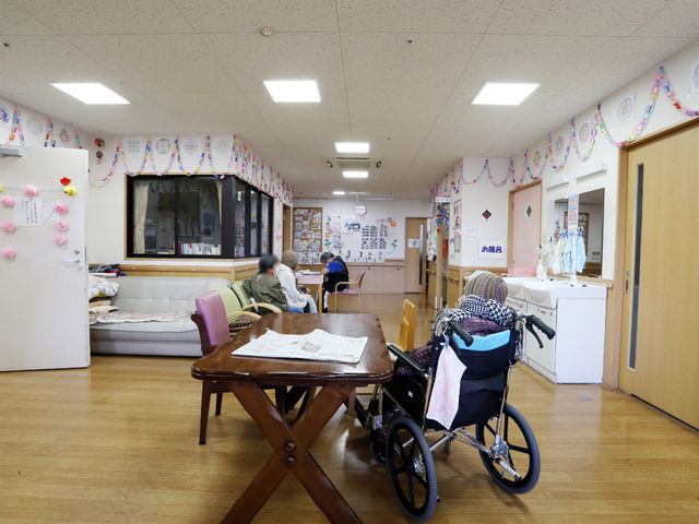 共有リビング空間 輪飾りで装飾された部屋 ゆったりとした共有リビングには、座ってくつろげるソファや個人の車椅子があり、暖かな雰囲気が感じられます。壁に飾られた色とりどりの飾りや掲示物も、明るい印象を与えています。