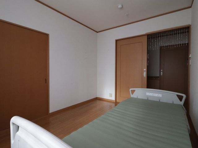 居室の寝具とドア 居室 広々とした居室は、落ち着いた色合いで統一され、快適な休息が取れる雰囲気です。ベッド横のスペースも十分で、動線を考えたつくりになっています。