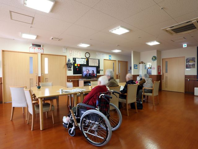 共有スペースの日常風景 食堂2 暖かい木目の共有スペースで、利用者様がテレビを見ながらくつろいでいます。明るく開放的なこの場所は、日々の憩いの場としてもご活用いただけます。