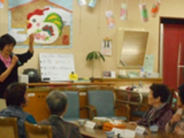 楽しげな和やかな空間 多目的室 スタッフが笑顔で手を挙げ、利用者の皆さんがゆったりと座りながら見守っている居心地の良い空間です。