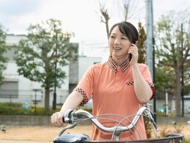 屋外で電話する職員 外観   屋外で自転車にまたがり、電話しながら笑顔を見せる職員の様子です。明るい雰囲気で、働く場の印象が伝わります。