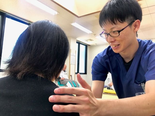 スタッフと利用者の機能訓練室 機能訓練室 笑顔で対応するスタッフが、座っている方の肩に手を添えて指導しています。安心して日々のリハビリが受けられる空間です。