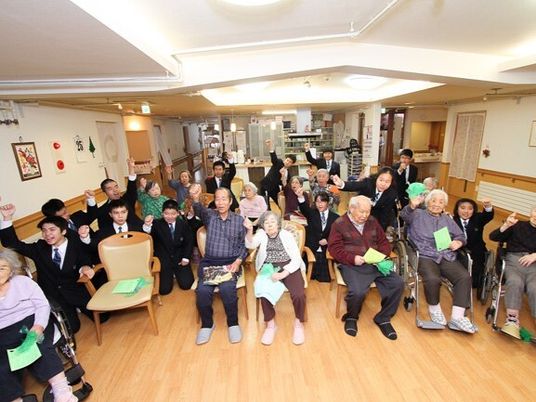スーツ姿の若者たちに囲まれて片手を上げるポーズをしている高齢者たち。共有スペースで。