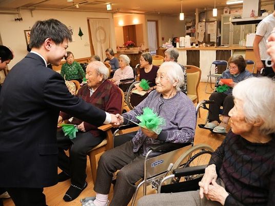 車いすの高齢女性が笑顔でスーツ姿の男性と握手をしている。手には緑の何かを持っている。