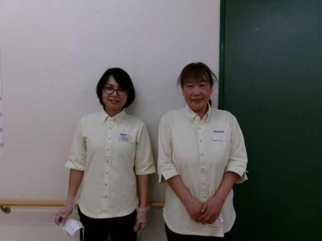 スタッフ2人の壁前 スタッフイメージ   壁の前で2人のスタッフが並んでいます。明るい色の制服姿で、やわらかな表情が印象的です。