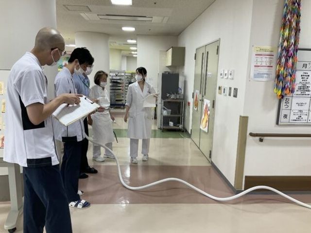 スタッフが廊下で訓練する様子 廊下   スタッフの方々が廊下に集まり、ホースを使った訓練を行っています。動線が広く確保されており、連携確認の場面が伝わります。