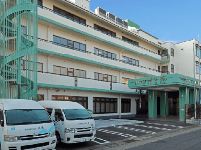 施設外観 外観   青空の下に、淡い色合いの建物が並ぶ外観です。送迎車も停まっており、出入りのしやすさがうかがえます。
