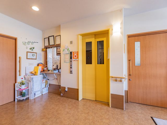黄色いエレベーターと廊下 エレベーター 手すり付きの廊下には温かみのある装飾が施され、心地よい空間が広がっています。黄色いエレベーターは明るさをプラスしており、利用者の方々の移動をサポートします。