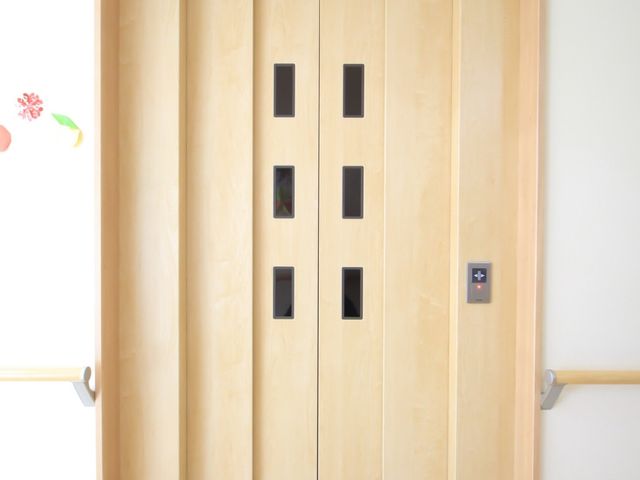 明るい木目調のエレベーター エレベーター ゆったりとした空間のエレベーターは、優しい木目調で落ち着いた雰囲気を演出しています。車椅子の方でも安心してご利用いただける様子を想像できます。
