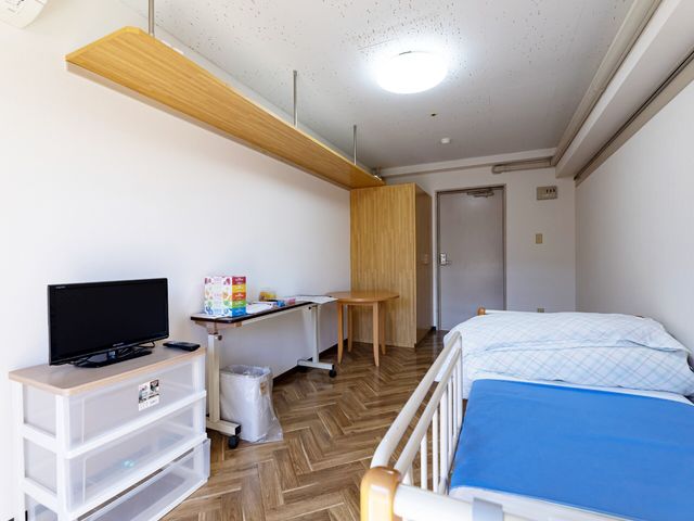 居室の入り口と家具 居室 ベッドとテレビがあり、ゆったりとした時間を過ごせる空間です。清潔感のある室内が印象的です。