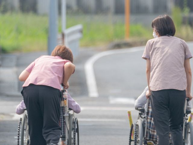 道を進む車椅子利用者とスタッフ 外観 スタッフが車椅子を押し、外部での散歩を楽しむ様子です。親しみやすい雰囲気が魅力です。