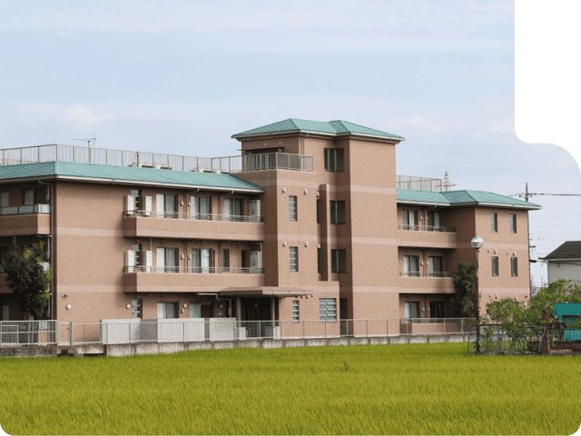 介護施設の外観 外観   緑の広がる前景に、落ち着いた色合いの建物が並んでいます。窓やベランダが多く、明るい暮らしを想像しやすい佇まいです。