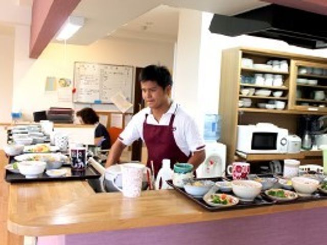 スタッフが立つダイニング 食事 笑顔で応対するスタッフがいる清潔感のあふれる食堂です。彩り豊かな料理で明るい食事時間を提供しています。