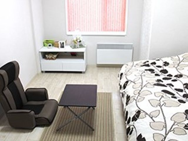 シンプルなプライベート居室 居室 落ち着いた色合いの居室で、ゆったりとした一人用の椅子が設置されています。明るい窓辺には小さな収納とデスクがあり、快適な個人空間が保たれています。