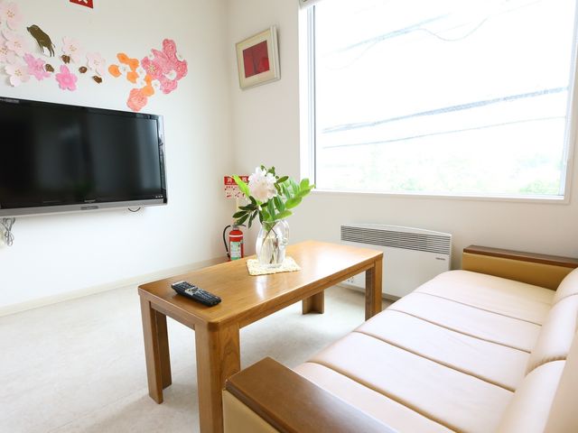 居室の明るい空間 共有スペース ゆとりある居室には、大型テレビとソファがあり、心地よい時間を過ごせる設計です。壁には花の装飾が施され、温かみのある環境が整っています。