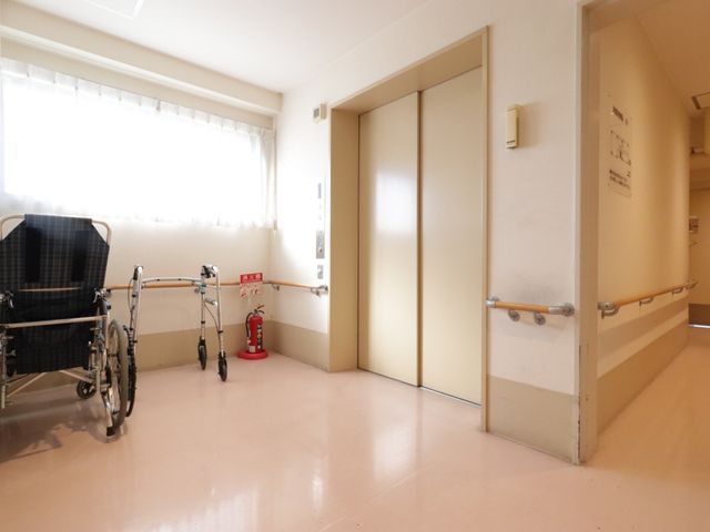 廊下の車椅子と歩行器 エレベーター 手すりが設置された明るい廊下です。車椅子や歩行器の横には、安全のための消火器も備えられています。